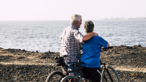 two seniors at rocky beach with their bikes - couple of pensioners hugged together looking at the sea or ocean - healthy and fitness lifestyle